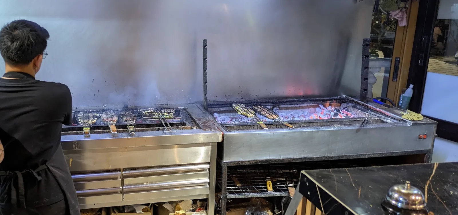 A cook at Grill Zone's long charcoal grill, fish baskets over glowing coals