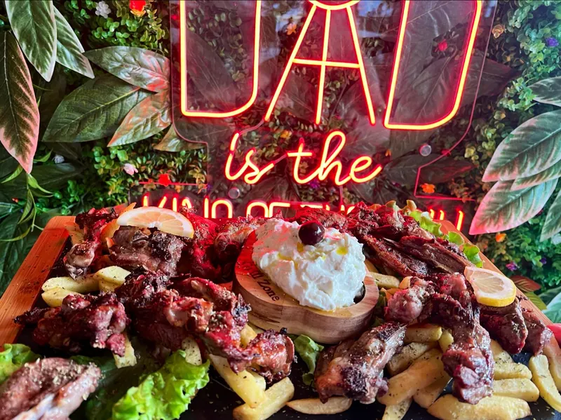 Grilled lamb and ribs on a wooden board with fries, lemon and tzatziki, under a red neon sign