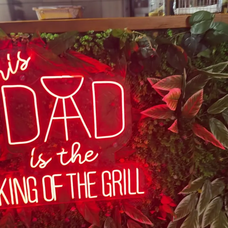 Red neon sign reading Dad is the king of the grill, on a wall of leaves