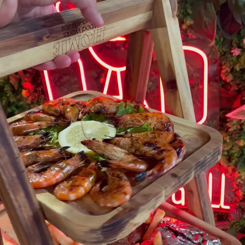 A tiered wooden stand of grilled prawns in front of the neon sign
